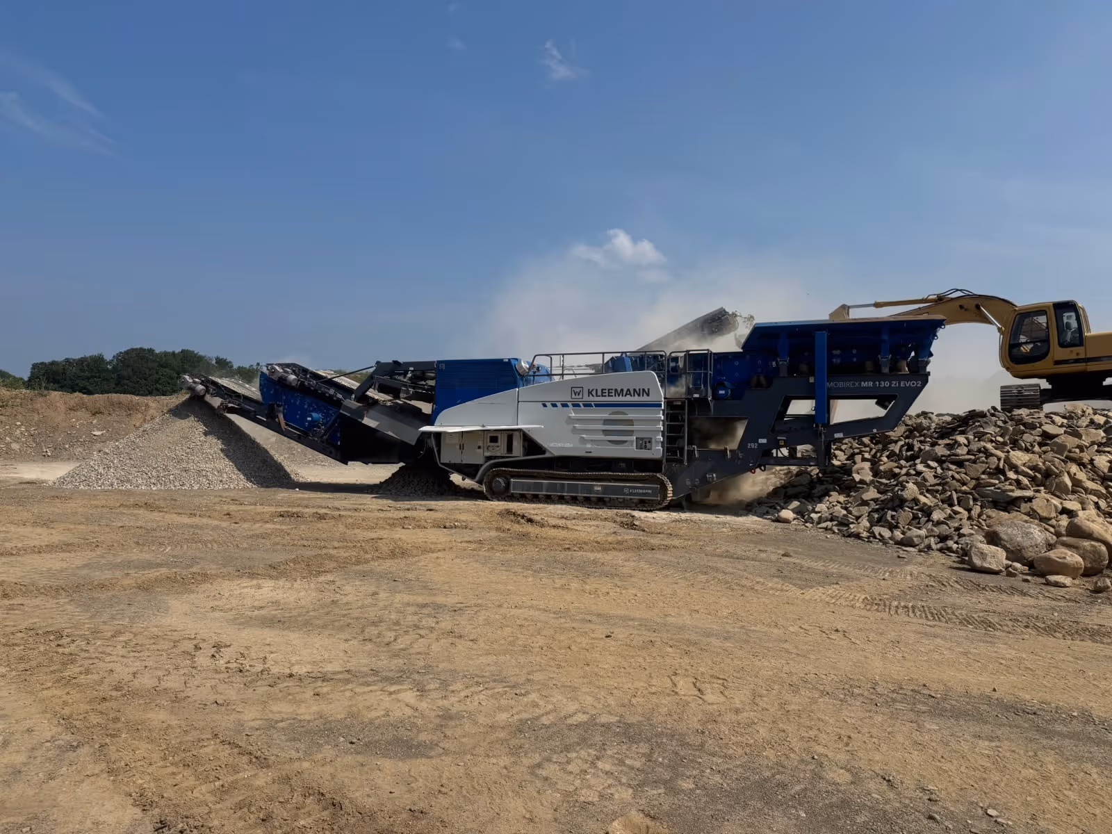 Kleemann mobile crusher processing rocks at a construction site with an excavator loading stones.