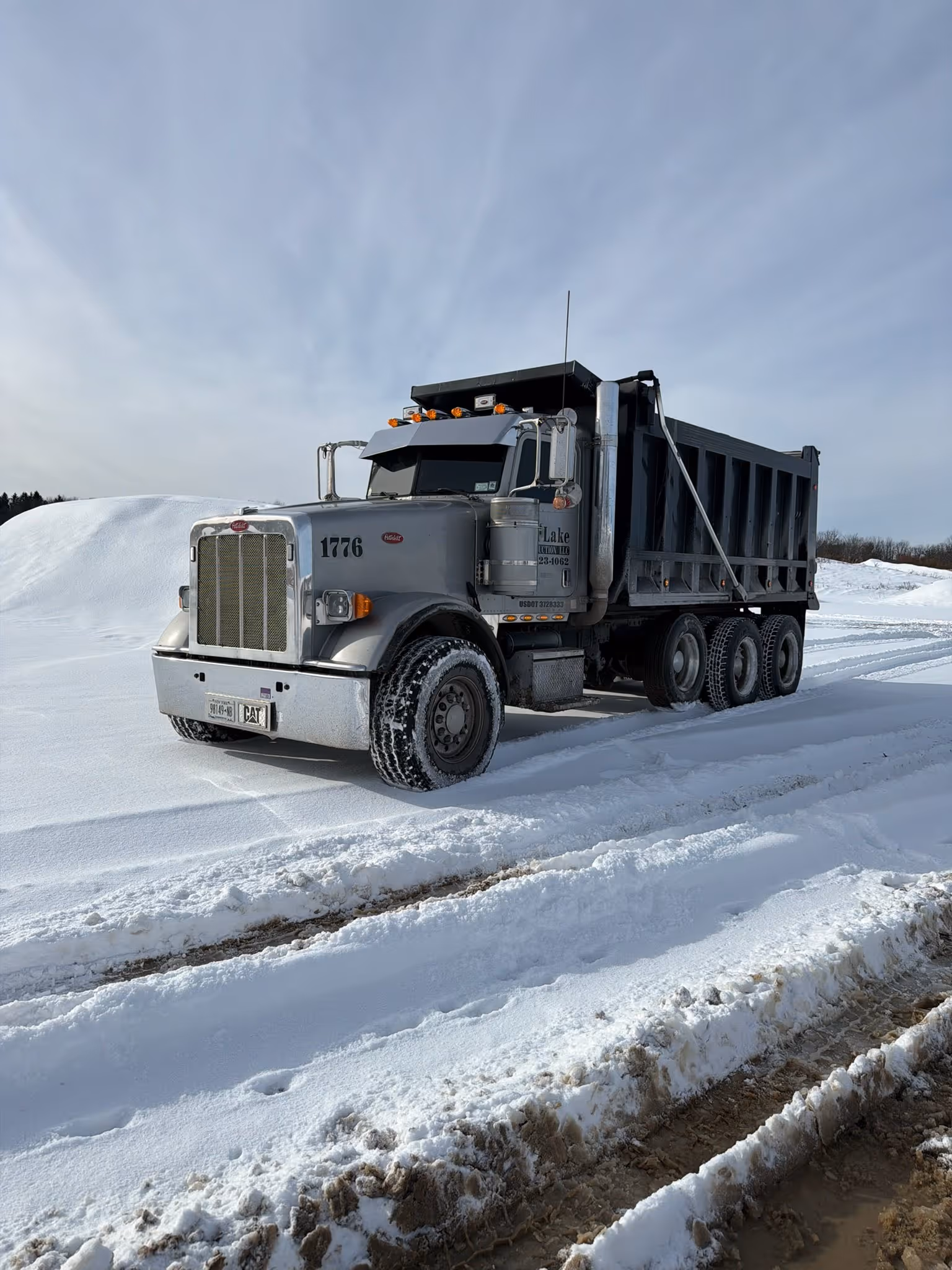 Silver dump truck with company logo parked on snow-covered ground with tire tracks and snow piles in the background.