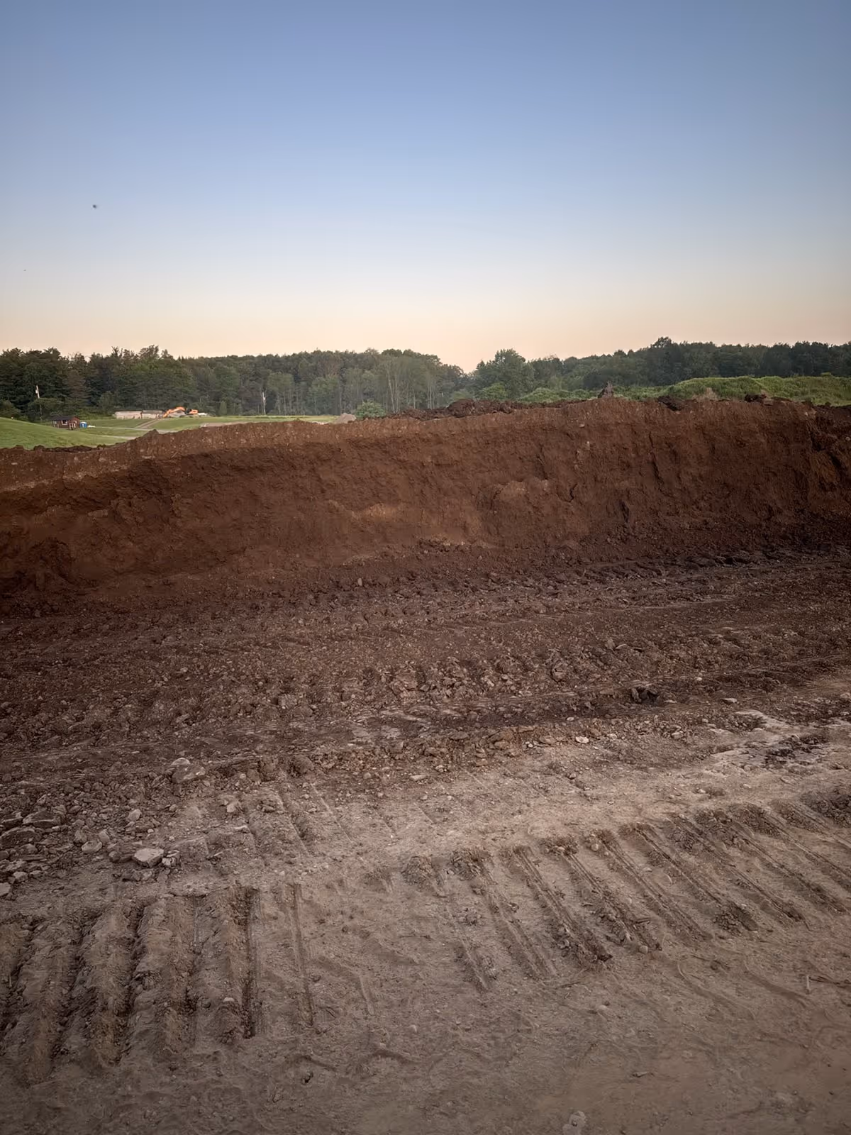 Large mound of freshly dug topsoil with tire tracks in the foreground and a tree-lined horizon under a clear sky.