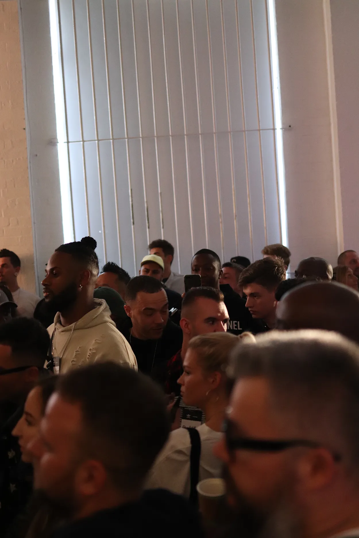 Crowded indoor gathering with people engaged in conversation near vertical blinds on a window.