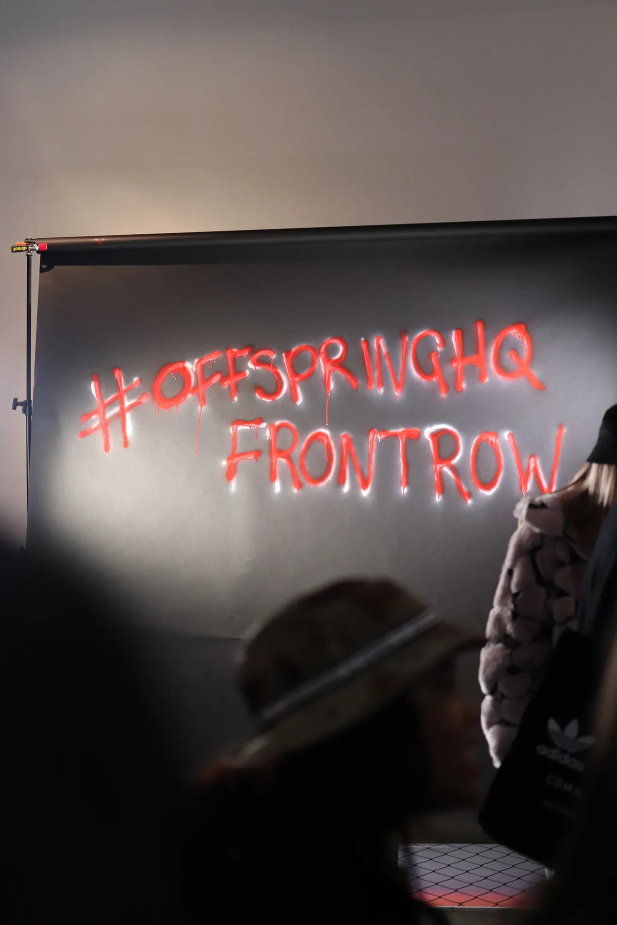 Black backdrop with red neon-style graffiti text reading '#OFFSPRINGHQ FRONTROW' with blurred people in front.