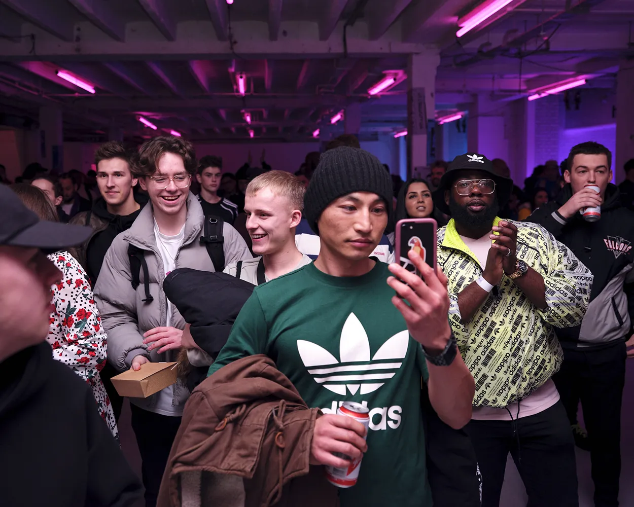 Young people standing in a purple-lit indoor space, one man in a green Adidas shirt looking at his phone and holding a drink can.