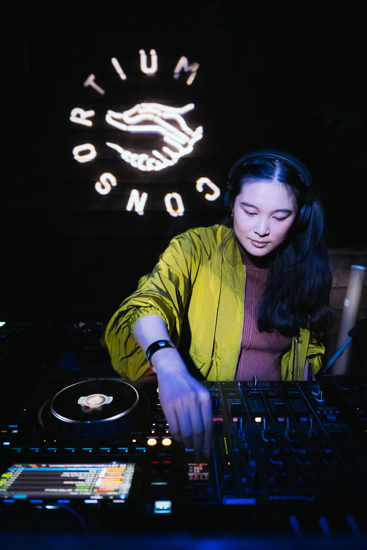 Female DJ wearing headphones and a yellow jacket adjusting controls on a DJ mixer in a dark room with Consortium logo on the wall behind.