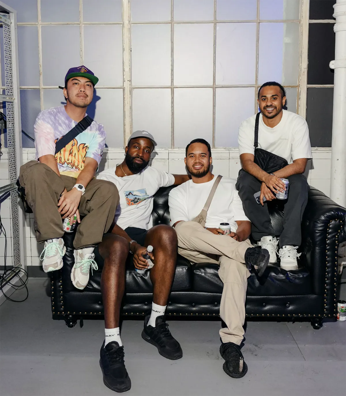Four men sitting closely on a black leather couch, holding drinks and smiling in a room with large frosted windows.
