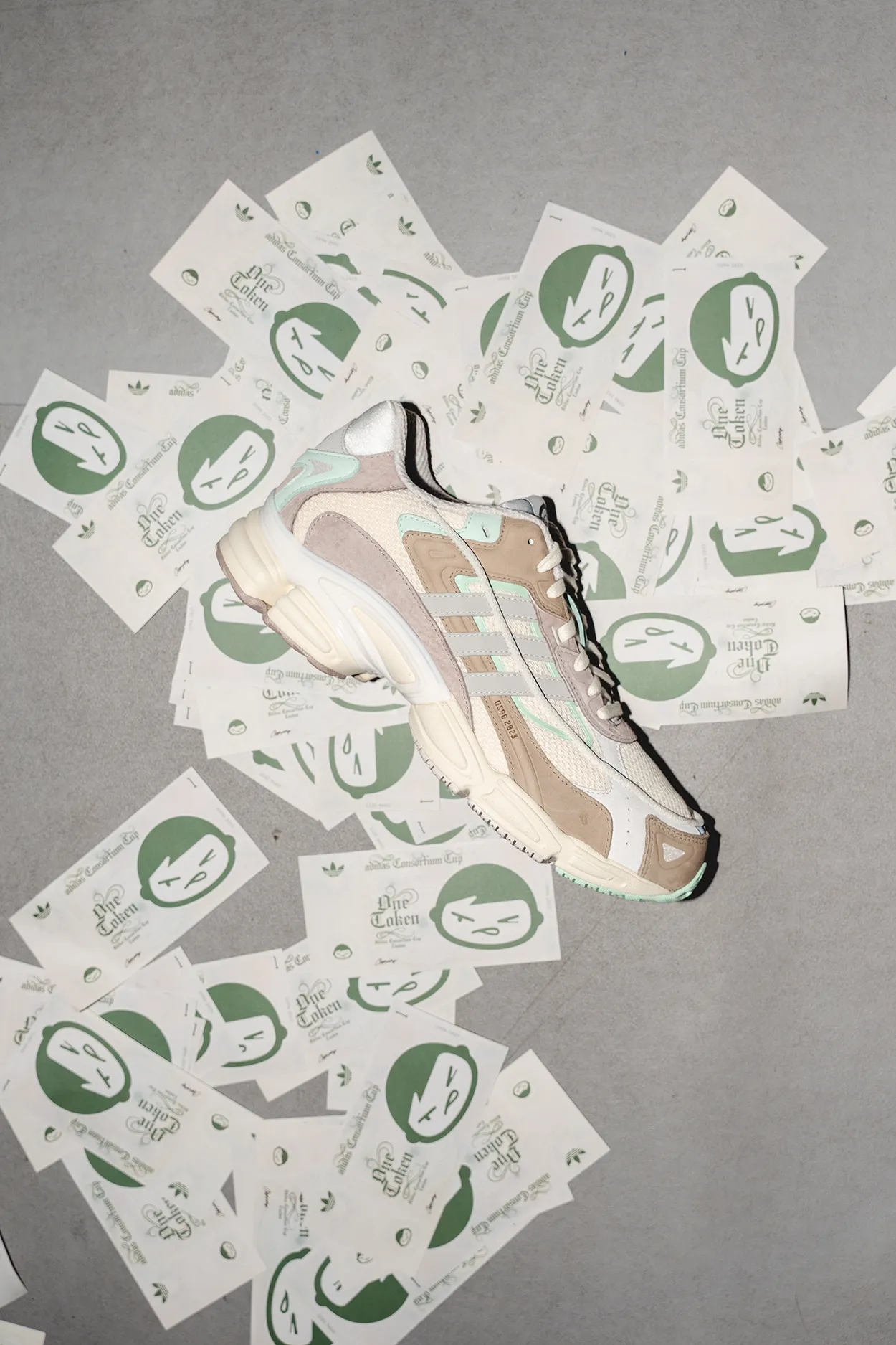 Beige and white adidas sneaker placed on a gray surface covered with scattered green and white stickers featuring a stylized face and text.
