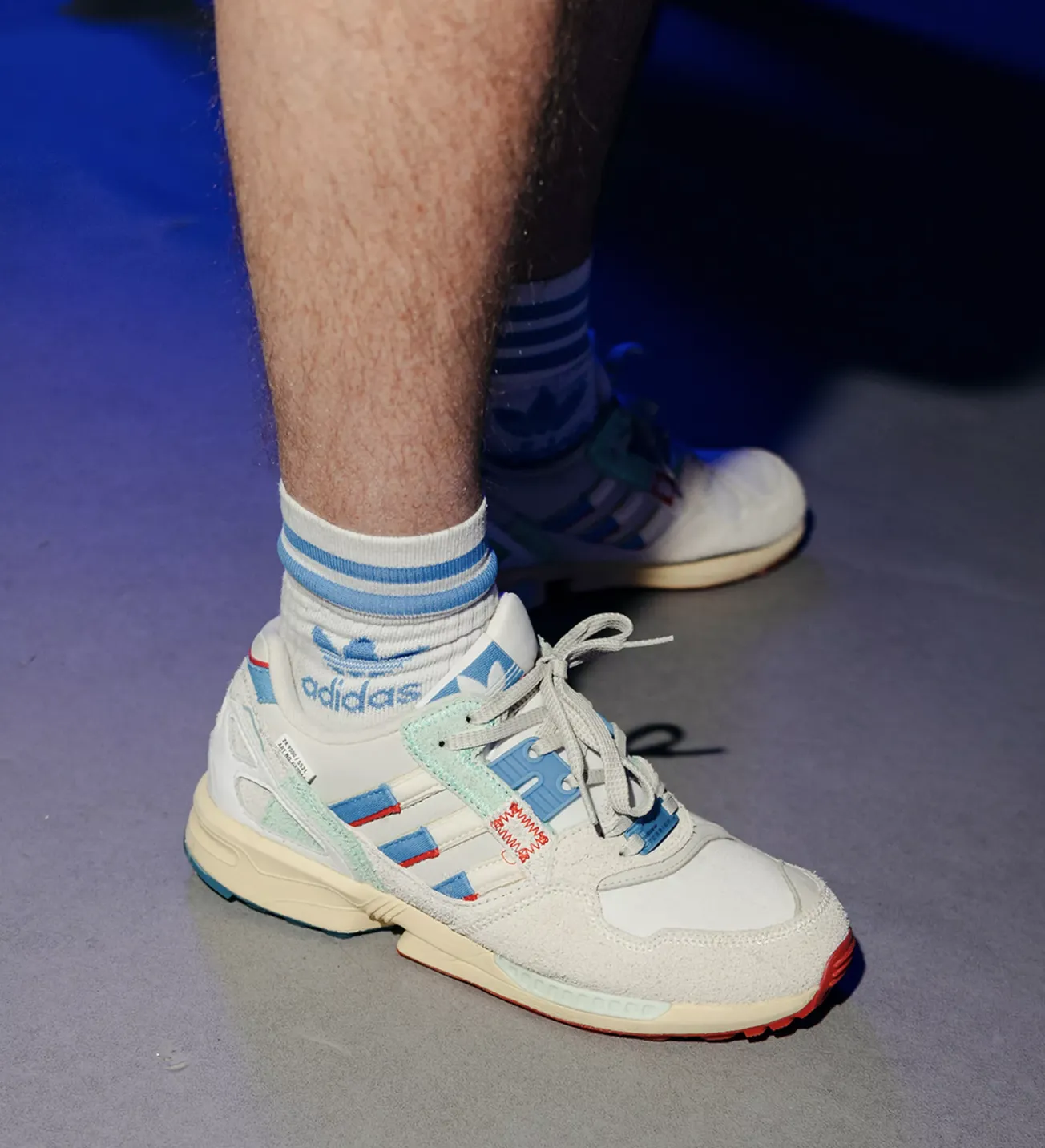 Person wearing white Adidas sneakers with blue and red accents and white Adidas socks with blue stripes.