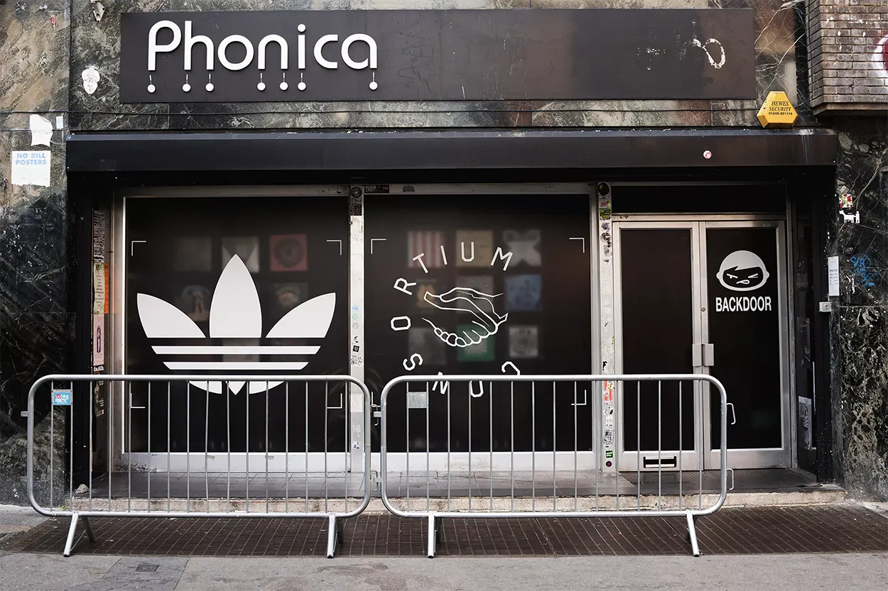 Storefront with black facade featuring large white Adidas trefoil logo, handshake graphic with 'Consortium' text, and 'Backdoor' logo with a stylized face, protected by metal barriers.