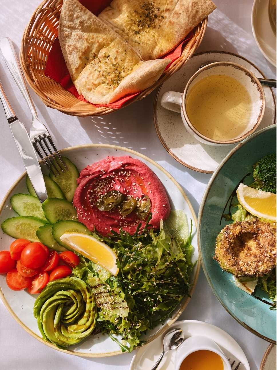 Teller mit rosa Hummus, Gurkenscheiben, Kirschtomaten, Avocadorose, Salat und Zitronenschnitz, daneben Brotkorb mit Fladenbrot und Tasse Kaffee.
