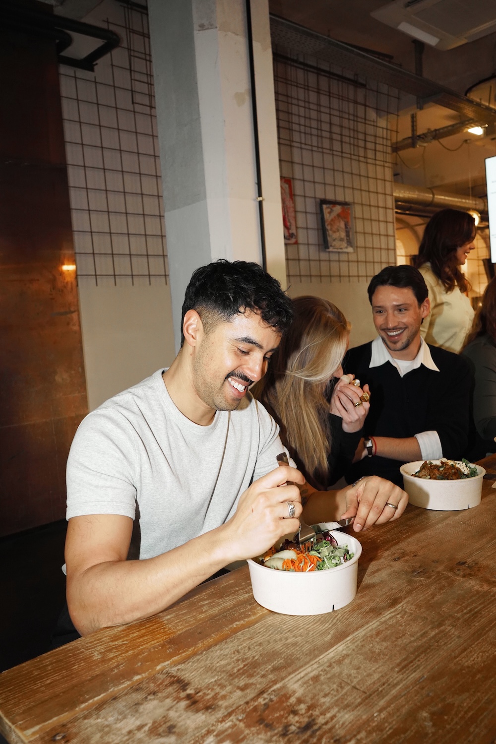 Junge Erwachsene sitzen an einem Holztisch in einem Restaurant und essen aus weißen Schalen, während sie lächeln und sich unterhalten.