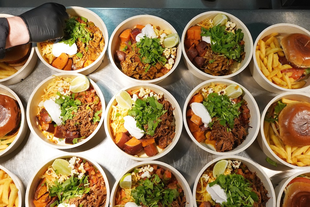 Mehrere Schalen mit gemischtem Essen aus Süßkartoffeln, Koriander, Sauerrahm und Limettenscheiben, daneben sind Burger mit Pommes.