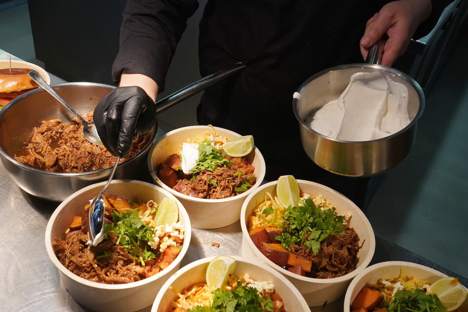 Person mit schwarzer Handschuh, der saure Sahne auf Schalen mit geschreddertem Fleisch, Mais, Limetten und Koriander verteilt.