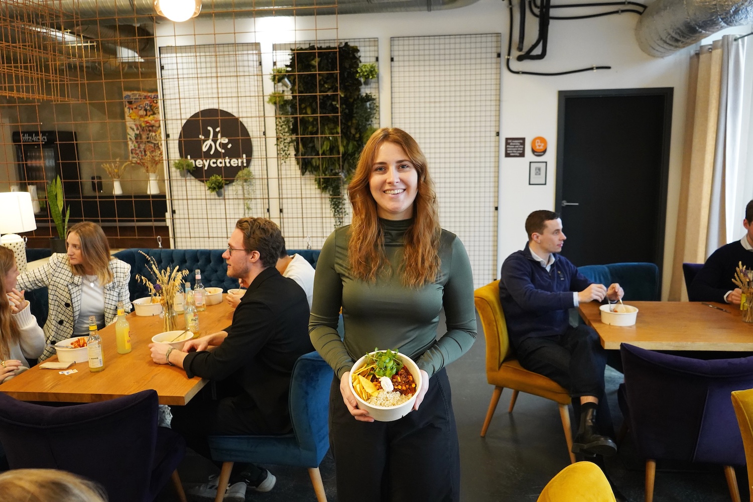 Frau mit langen roten Haaren hält eine Schüssel mit Essen und lächelt in einer modernen Cafeteria mit mehreren Personen im Hintergrund.