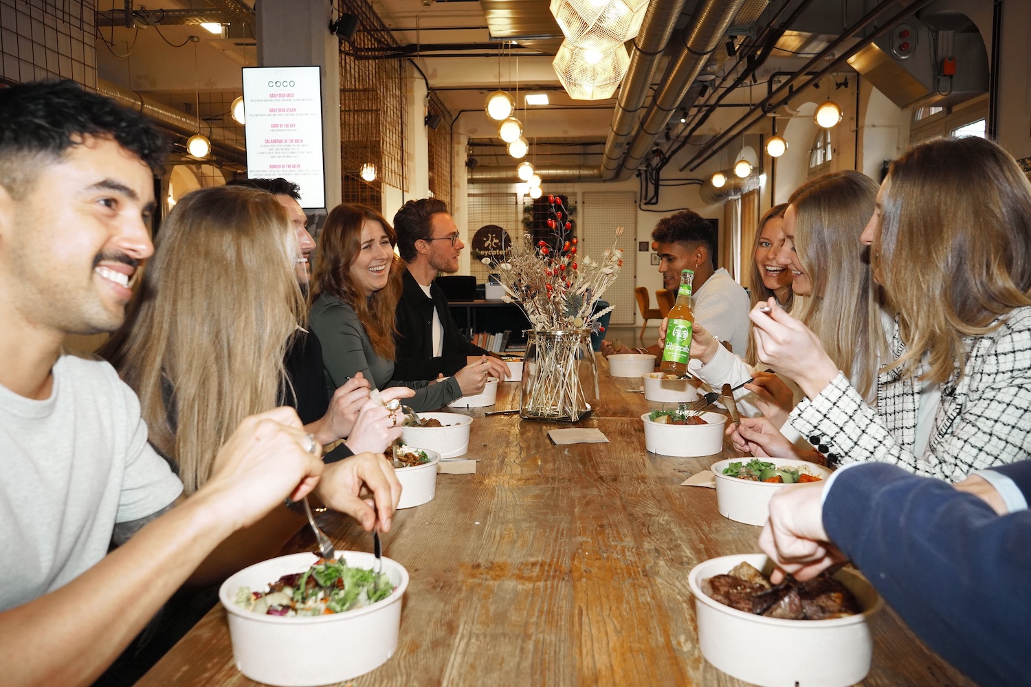 Gruppe Kollegen sitzt an einem langen Holztisch in einem modernen Restaurant und isst zusammen, während sie lachen und sich unterhalten.