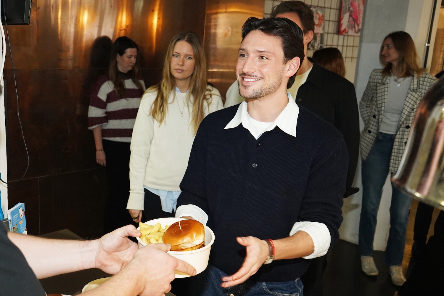 Ein lächelnder Mann im schwarzen Pullover nimmt einen Teller mit Burger und Pommes von einer anderen Person entgegen, im Hintergrund stehen mehrere Menschen in einer Innenraumumgebung.