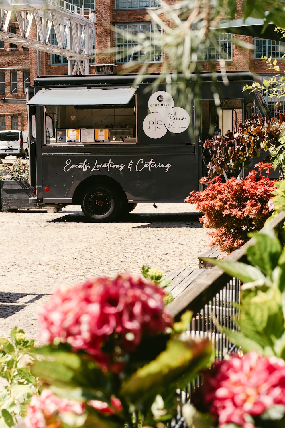 Schwarzer Food-Truck mit der Aufschrift ‚Events, Locations & Catering‘ steht auf einem gepflasterten Platz vor einem Backsteingebäude, umgeben von bunten Blumen.