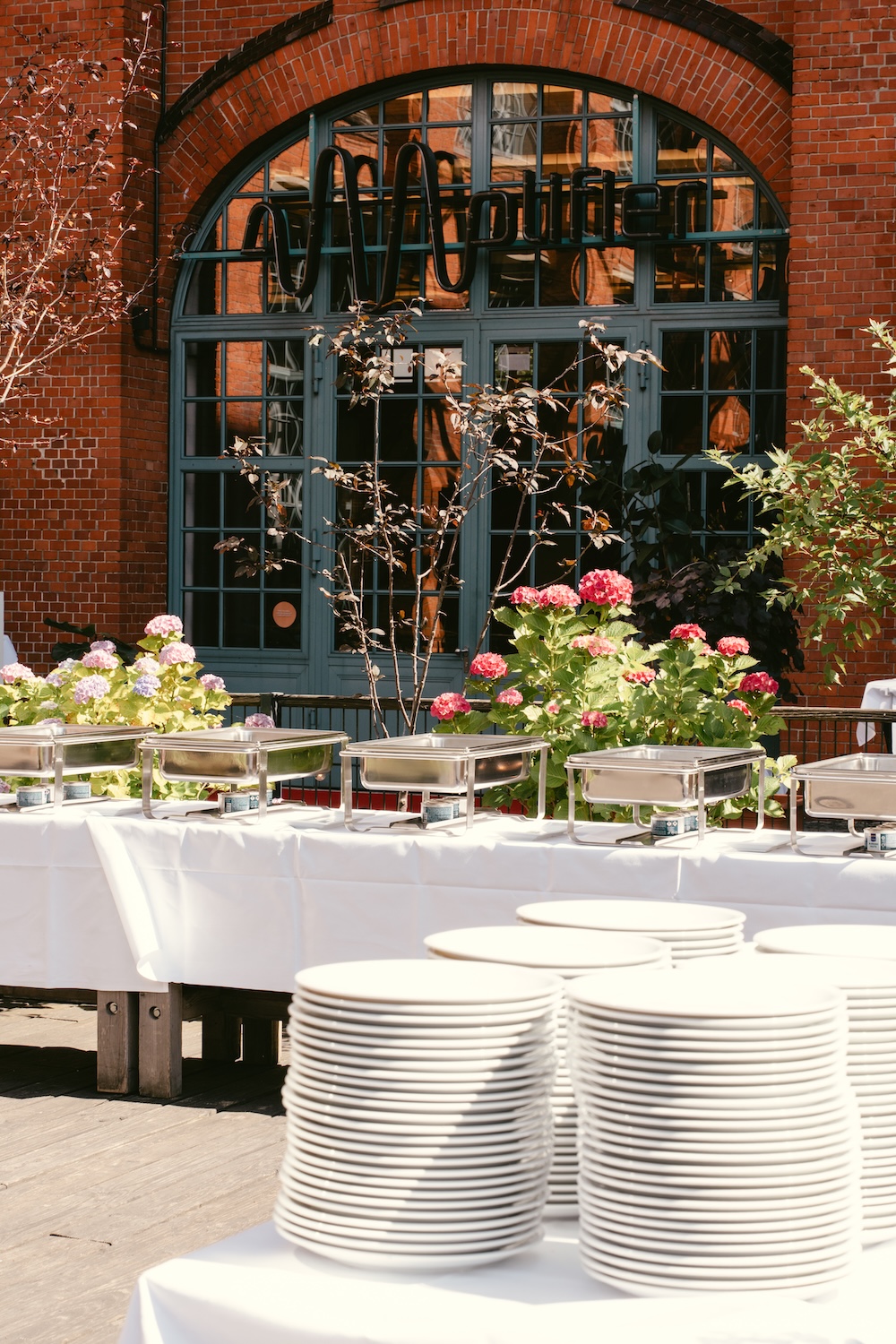 Buffet-Tisch mit leeren weißen Tellern und Blumen vor einem roten Backsteingebäude mit einem großen Fenster.