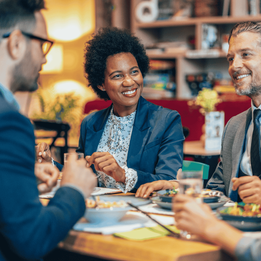 Drei beruflich gekleidete Personen, die bei einem Essen an einem Tisch miteinander lachen und sich unterhalten.