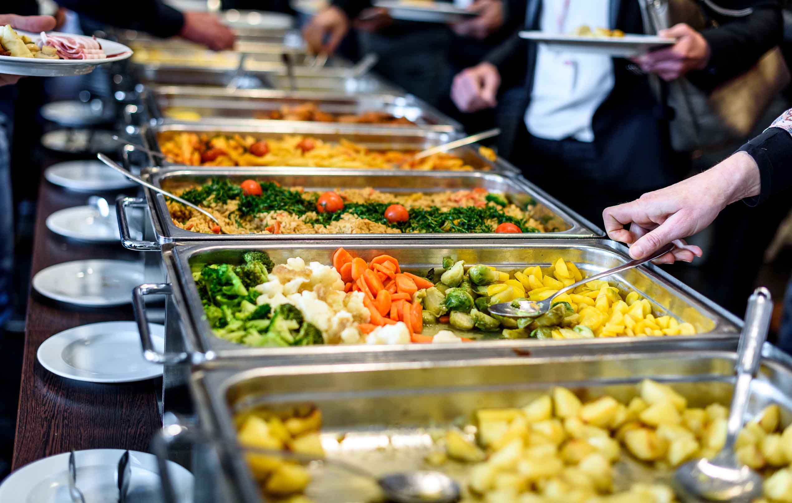 Buffet mit verschiedenen warmen Speisen wie Gemüse, Reis mit Tomaten und Kartoffeln, Menschen bedienen sich.