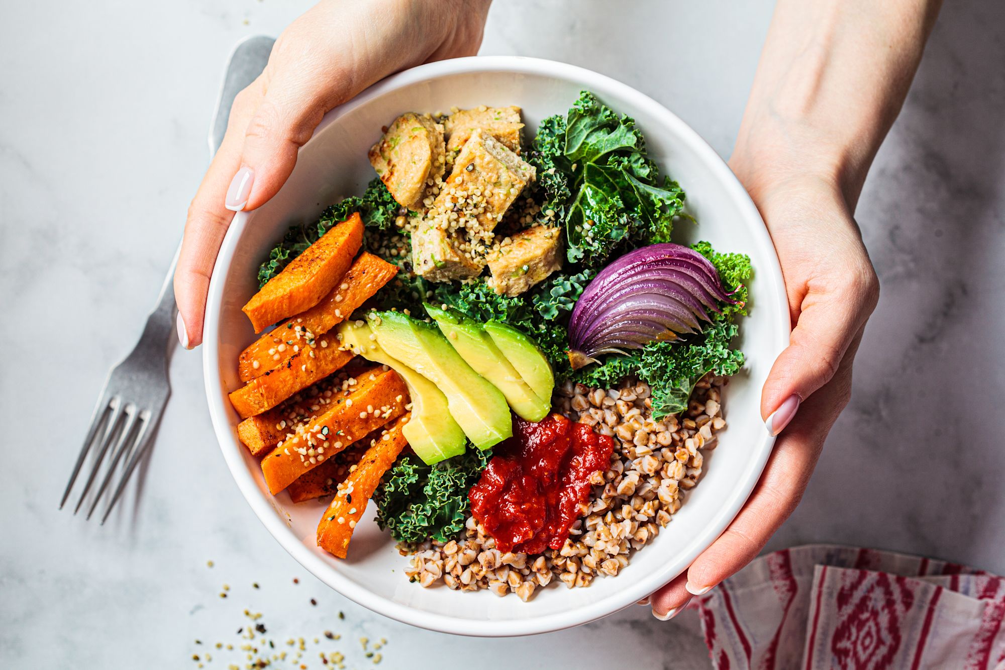 Vier lachende junge Erwachsene, zwei halten eine bunte Schüssel mit Salat und Falafel vor einem orangefarbenen Hintergrund.