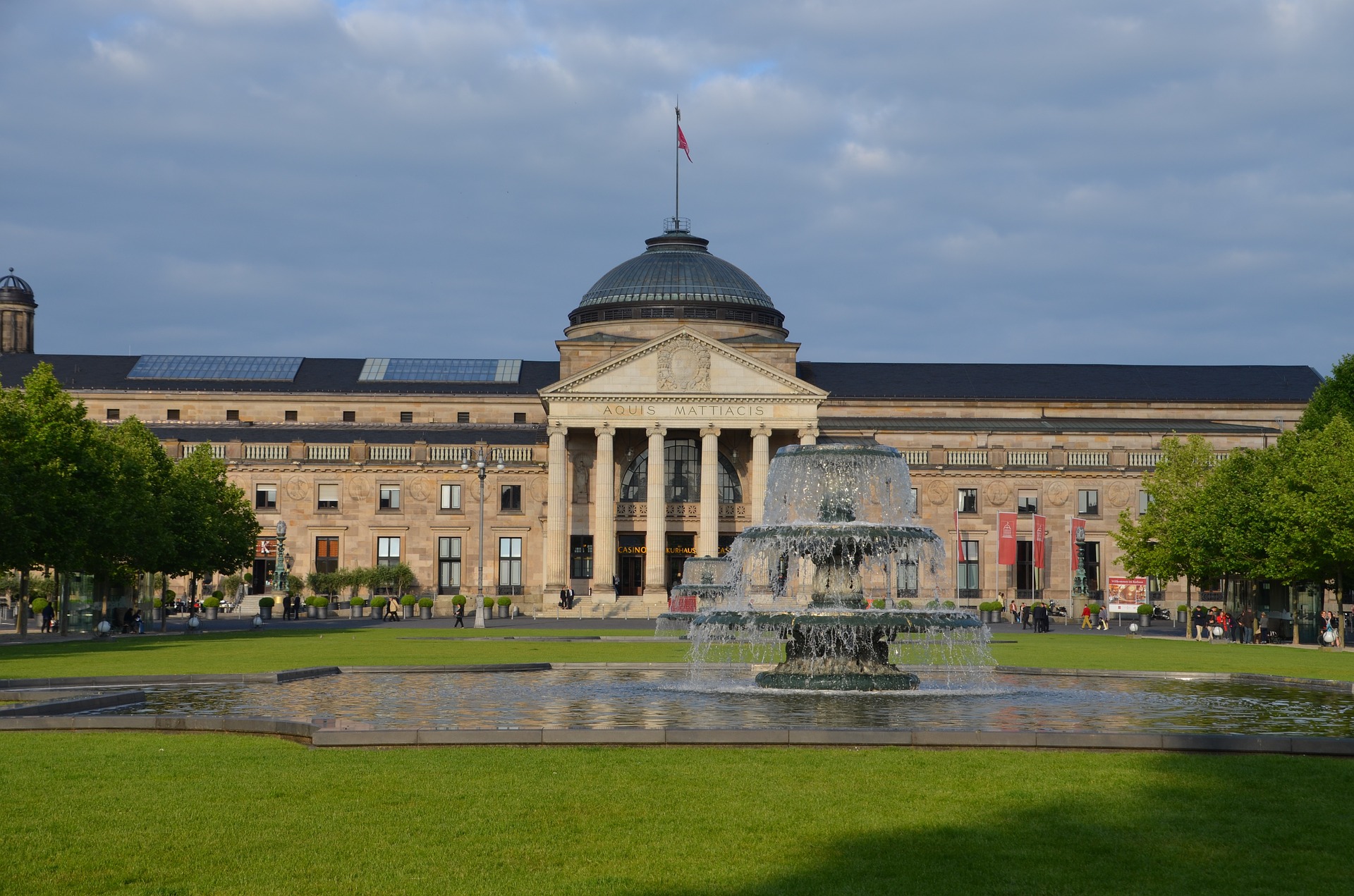 Vier lachende junge Erwachsene, zwei halten eine bunte Schüssel mit Salat und Falafel vor einem orangefarbenen Hintergrund.
