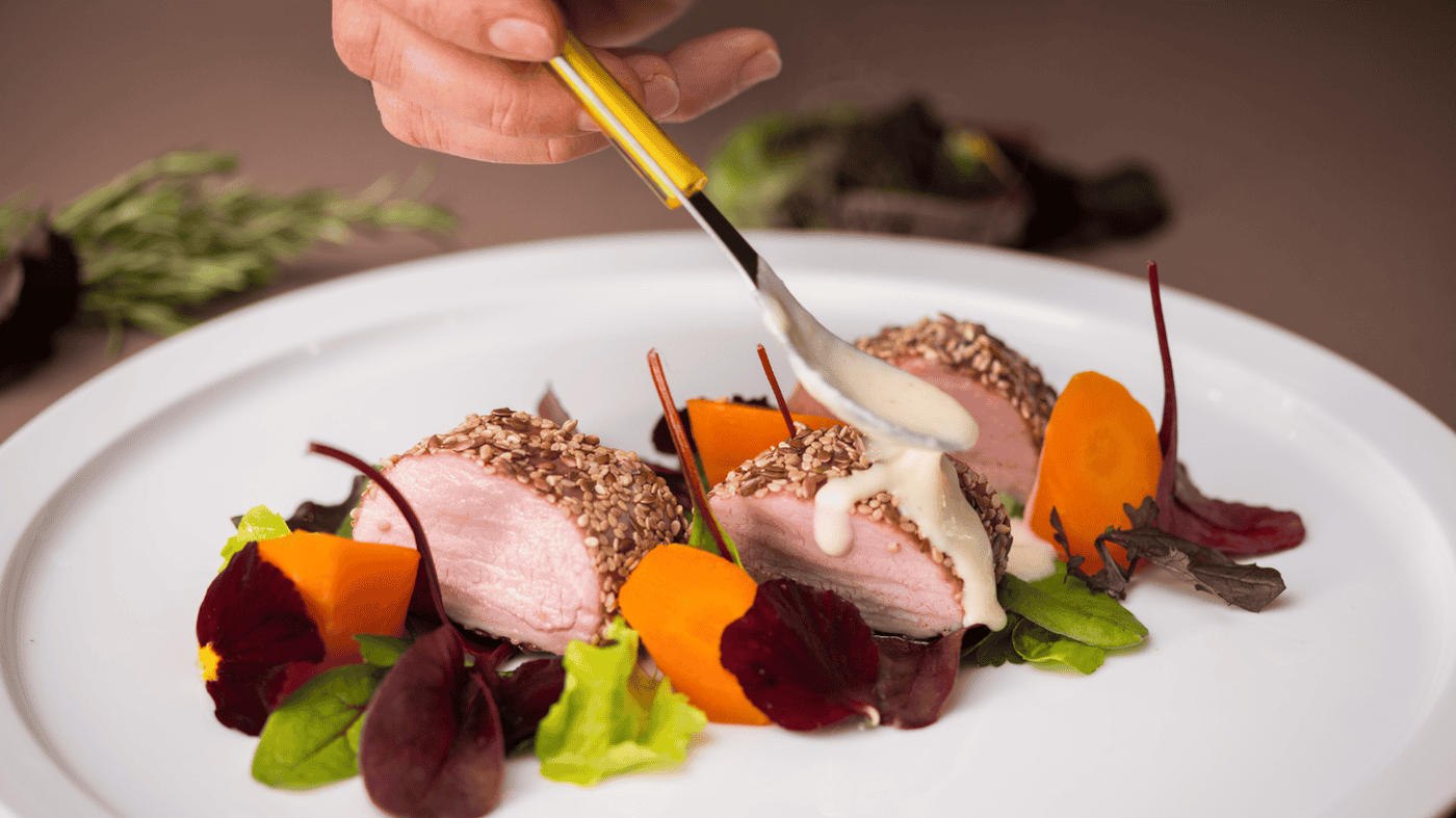 Vier lachende junge Erwachsene, zwei halten eine bunte Schüssel mit Salat und Falafel vor einem orangefarbenen Hintergrund.