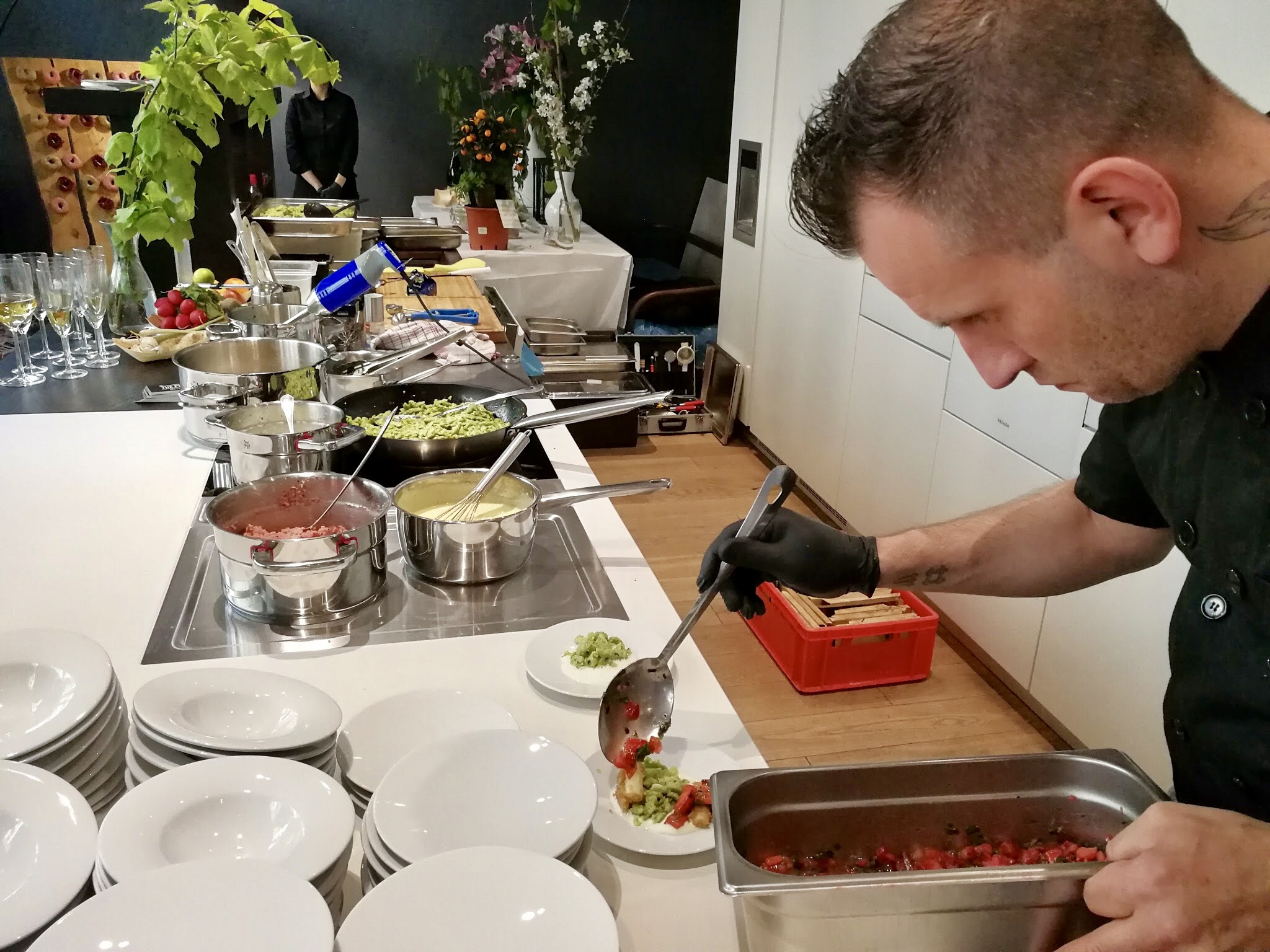 Vier lachende junge Erwachsene, zwei halten eine bunte Schüssel mit Salat und Falafel vor einem orangefarbenen Hintergrund.