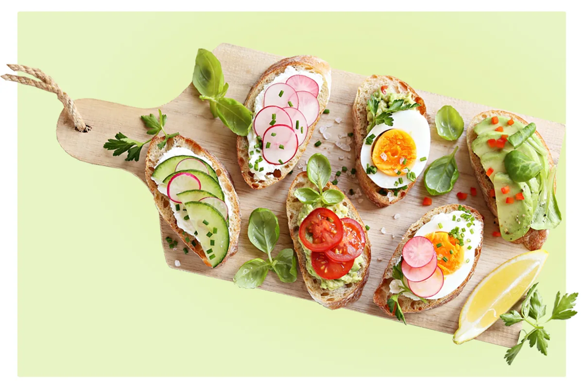 Four laughing young adults, two holding a colorful bowl of salad and falafel against an orange background.