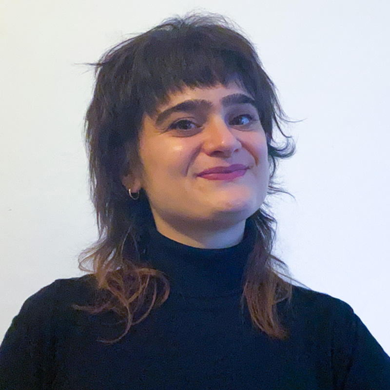 Smiling person with short layered hair and bangs wearing a black turtleneck against a plain light background.