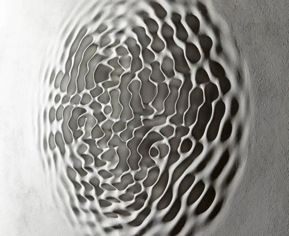 Cymatics sound-wave ripple pattern formed in a grey plaster surface.