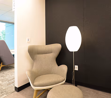 Tan upholstered lounge chair and ottoman beside a glowing floor lamp against a dark acoustic felt wall.