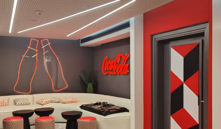 Coca-Cola branded breakout lounge with acoustic ceiling and neon bottle wall art.