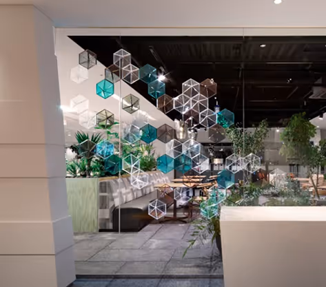 Restaurant interior divided by hanging translucent geometric acoustic modules with greenery.