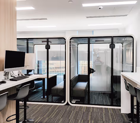 Open-plan office with two black-framed acoustic meeting pods beside desks by tall windows.