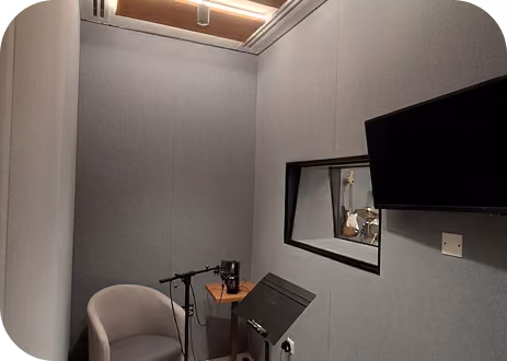 Acoustic recording booth with grey fabric wall panels, microphone, armchair and wall monitor.