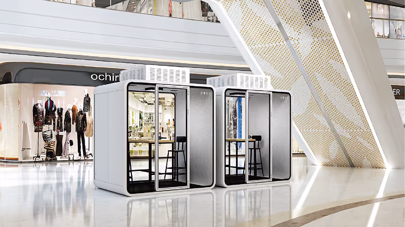 Two white soundproof PrimePod office booths with bar stools inside a modern shopping mall.