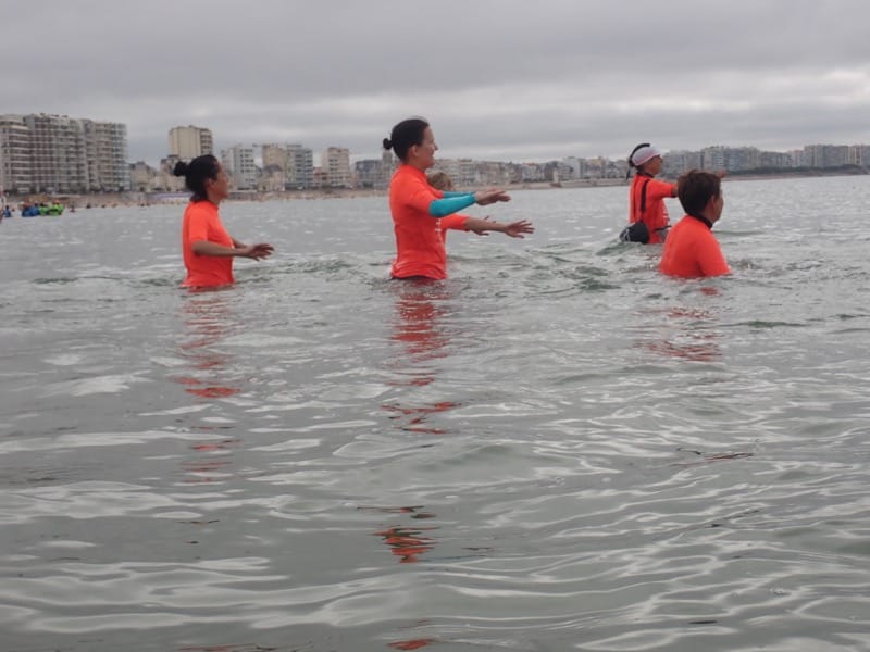 Four people wearing orange shirts standing waist-deep in calm sea water near a coastal city under an overcast sky.