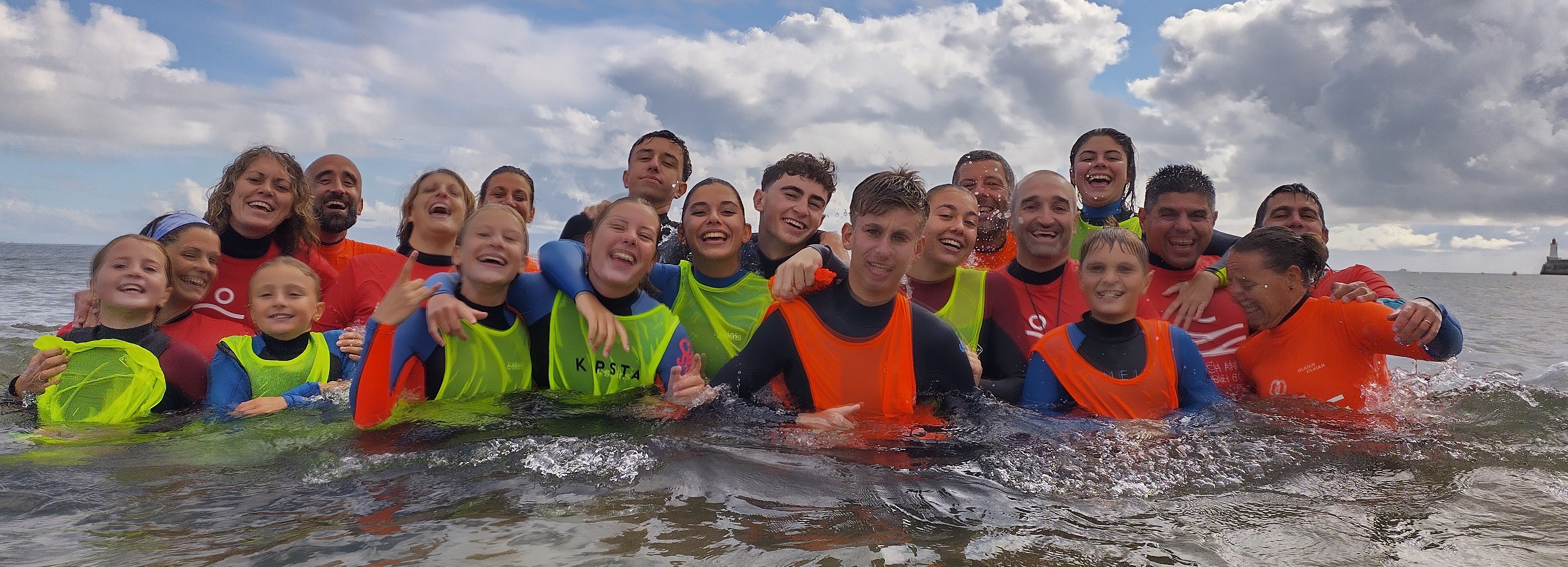 Group of smiling people wearing colorful wetsuits standing together in shallow ocean water under a partly cloudy sky.