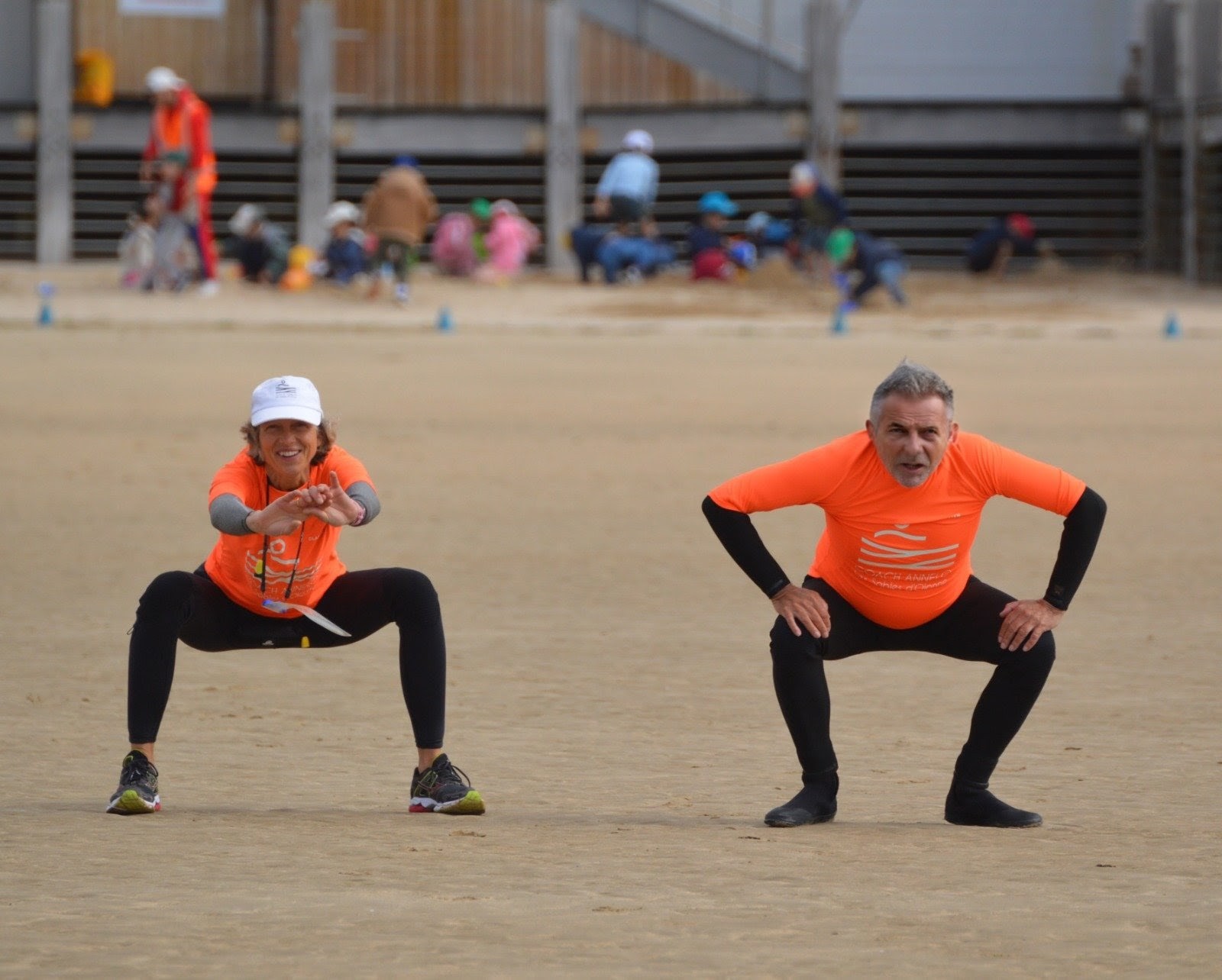 Two women in athletic wear doing squats and smiling on a sandy beach near the ocean.