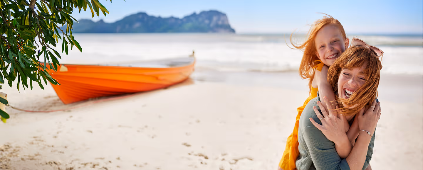 Smiling woman and child with red hair hugging on a sandy beach near an orange boat with a mountain in the background.