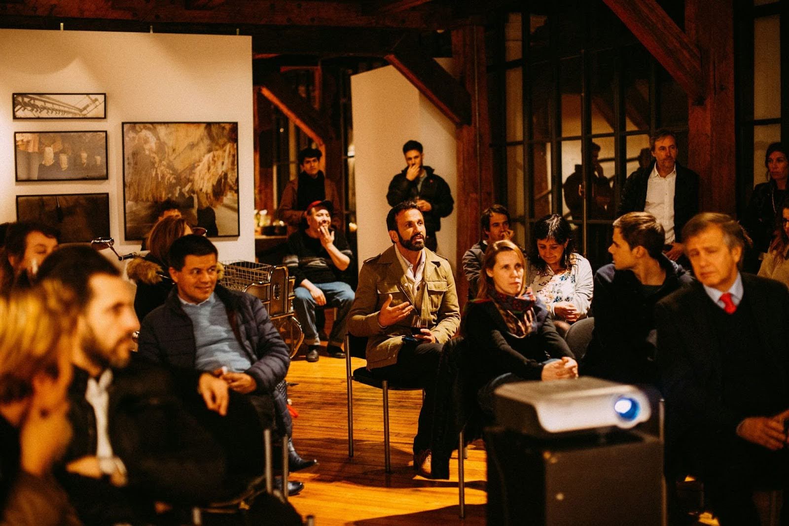 Audience seated indoors attentively listening, with a man in a tan jacket speaking, surrounded by warm wooden beams and framed artwork.