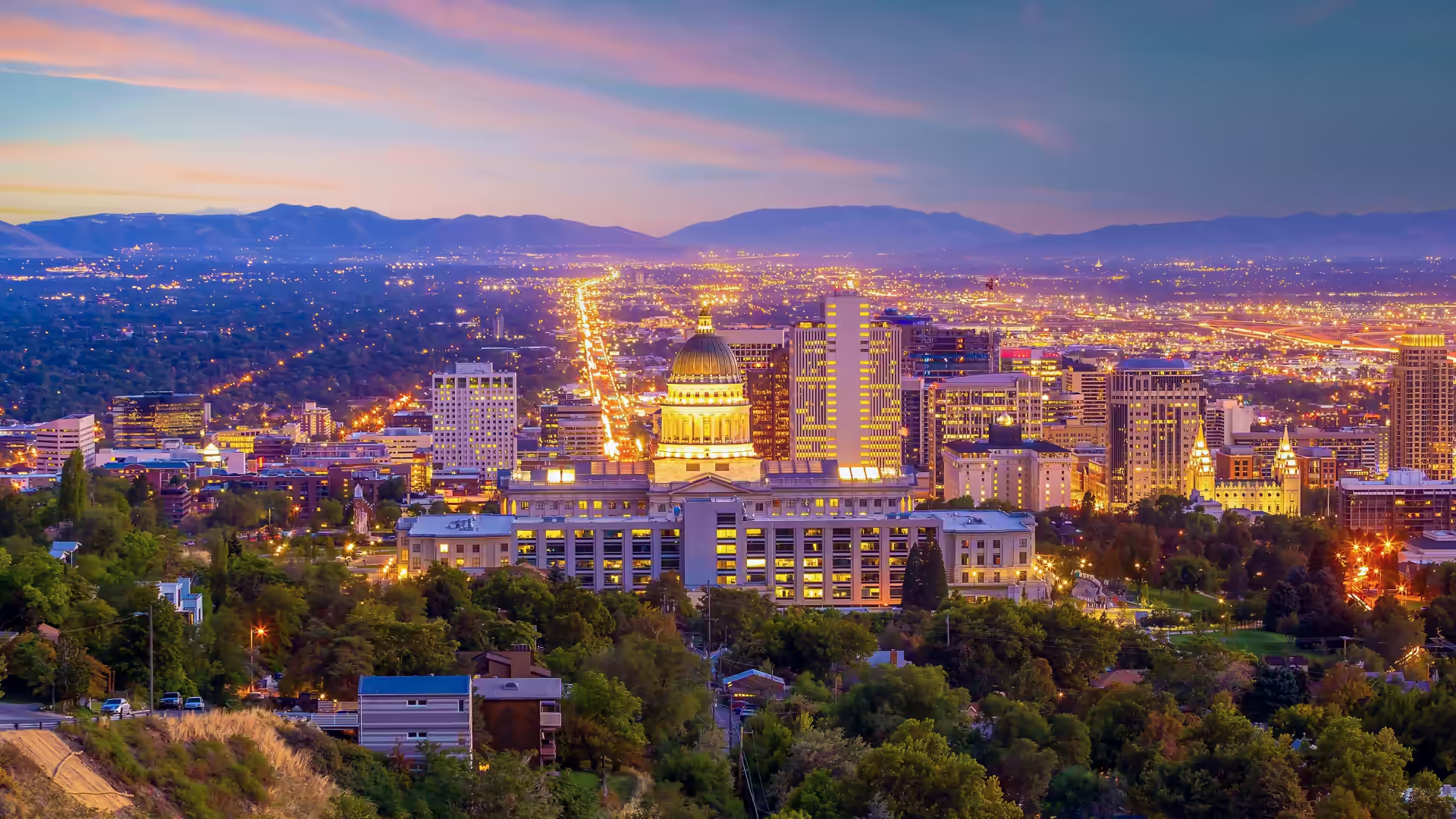 Night view of Salt Lake City with a prominent illuminated capitol building and surrounding skyscrapers against a backdrop of mountains and a colorful sky.