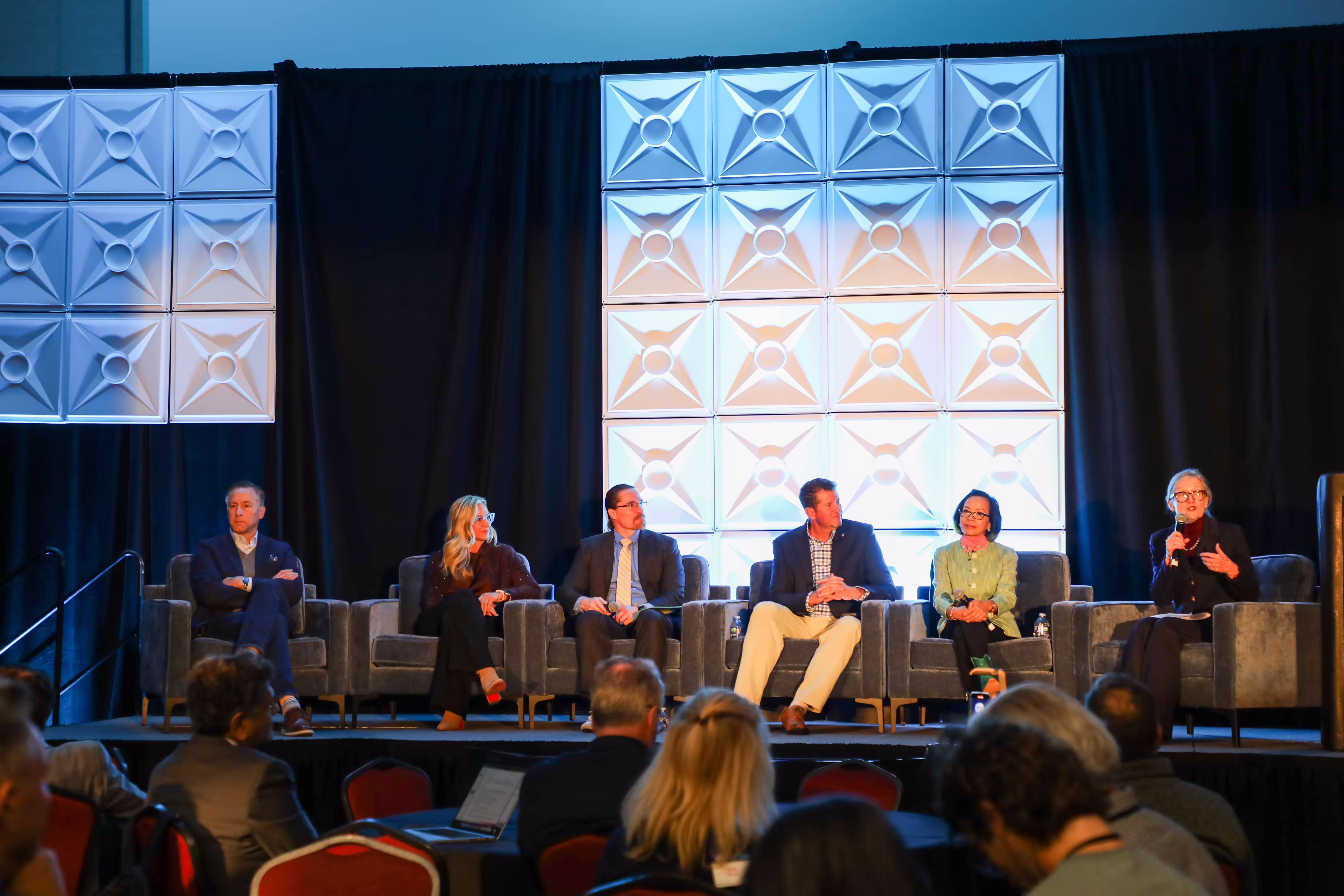 Panel of six speakers seated on stage with audience in foreground at a conference.