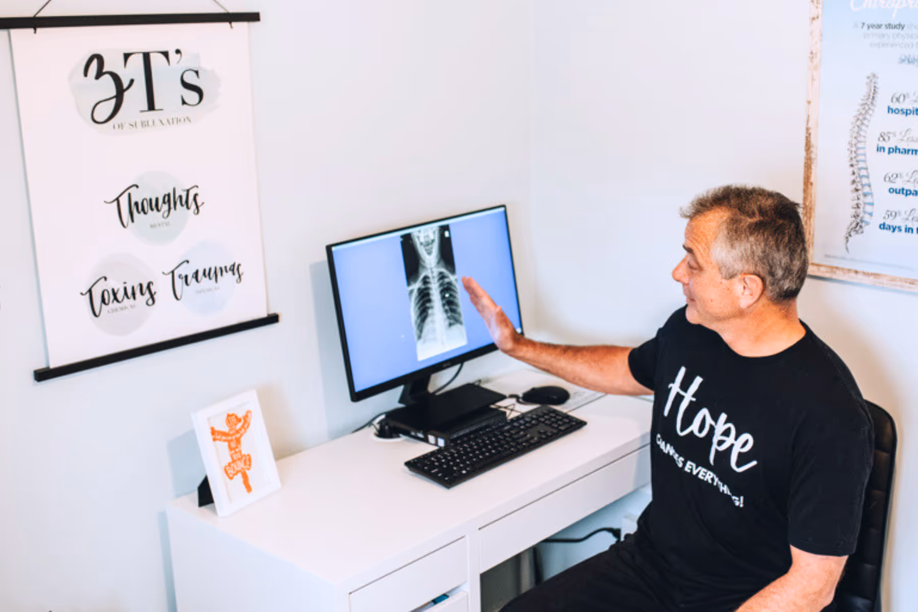 Man wearing a 'Hope Changes Everything' shirt pointing at an X-ray image of a chest displayed on a computer monitor in a medical office.