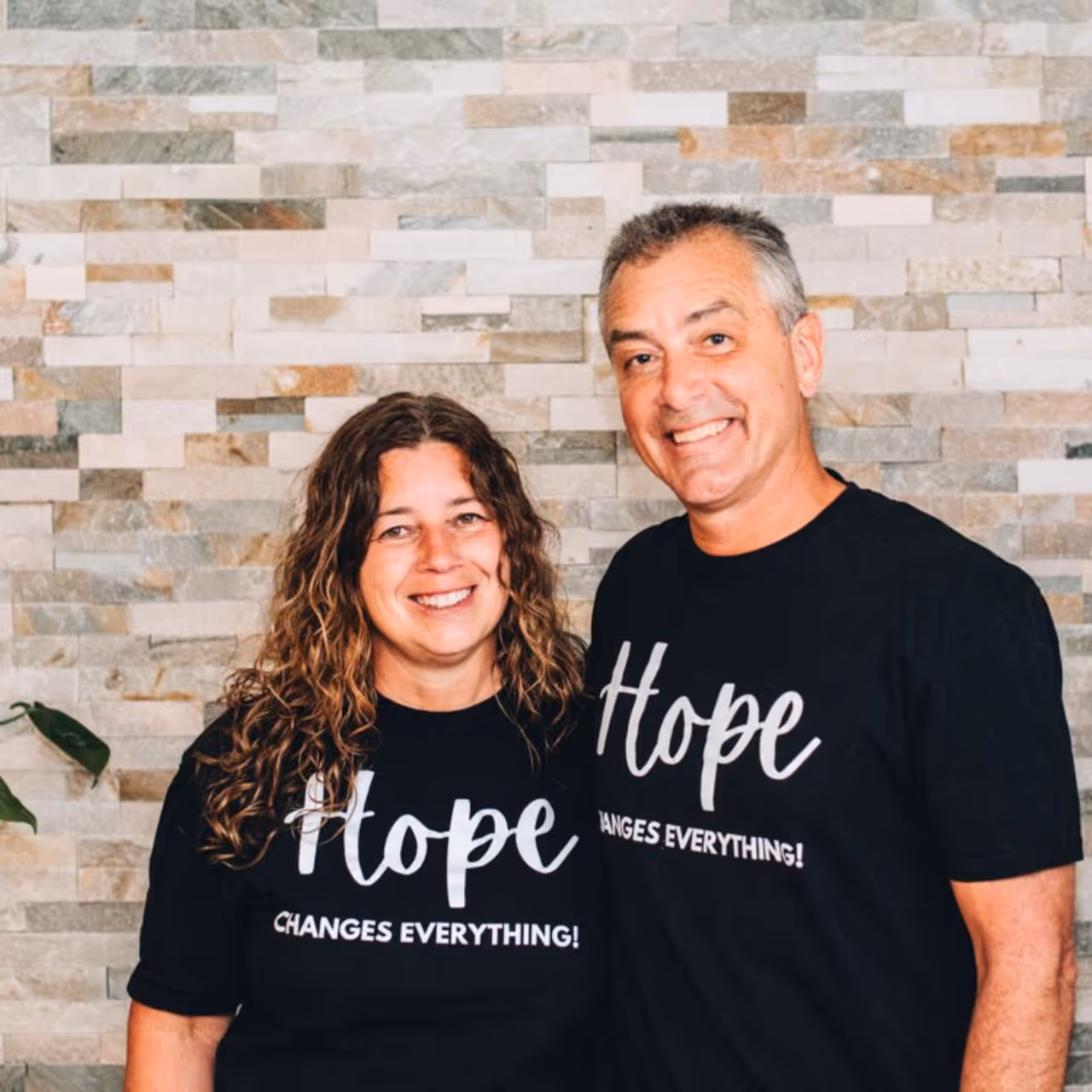 Three people smiling in front of a stone wall, wearing black shirts with motivational messages.
