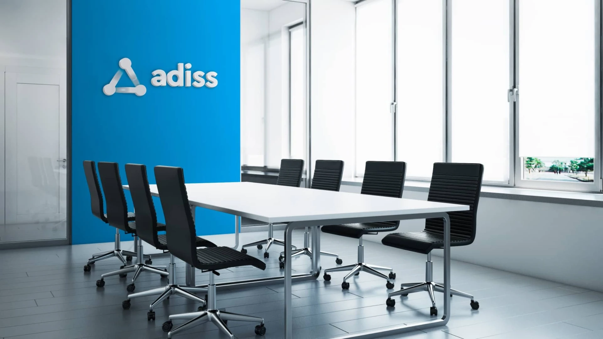 Interior de una sala de reuniones moderna con una mesa blanca rectangular rodeada de sillas negras, presentando una pared azul brillante al fondo con el logotipo de "adiss" en relieve blanco y grandes ventanales a la derecha