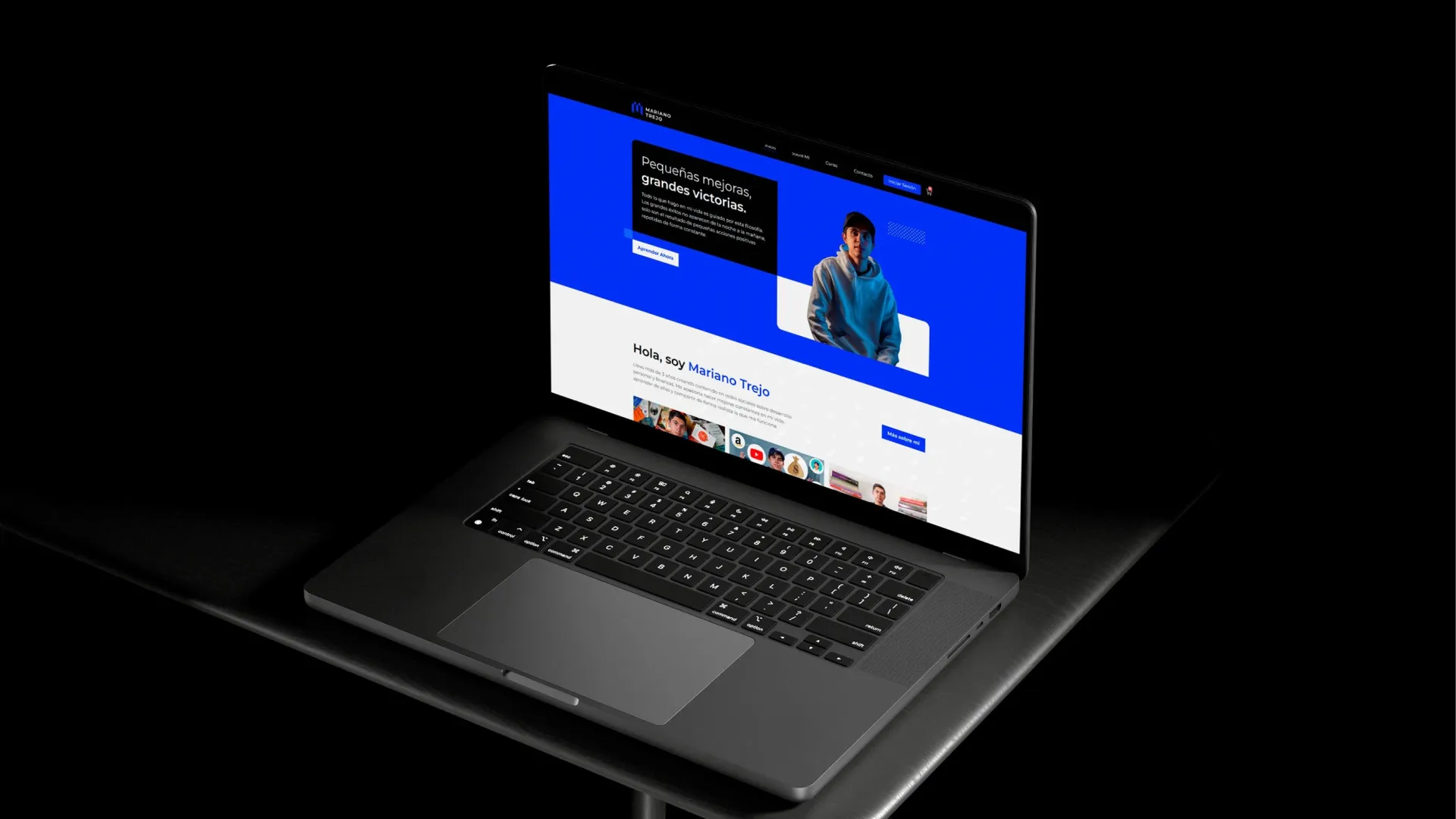 Computadora portátil negra abierta sobre una superficie oscura, mostrando en su pantalla una página web con diseño azul y blanco que incluye la frase 'Pequeñas mejoras, grandes victorias' y una fotografía de un joven