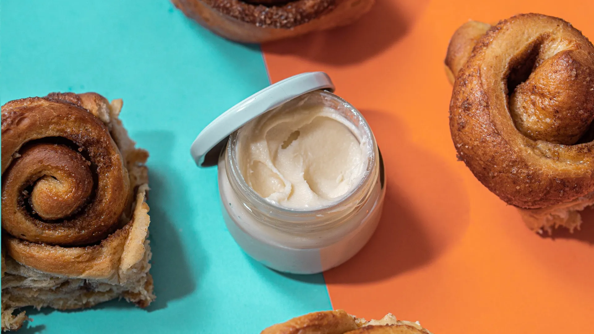 Rollos de canela acompañados de frasco con crema sobre fondo dividido en colores vibrantes