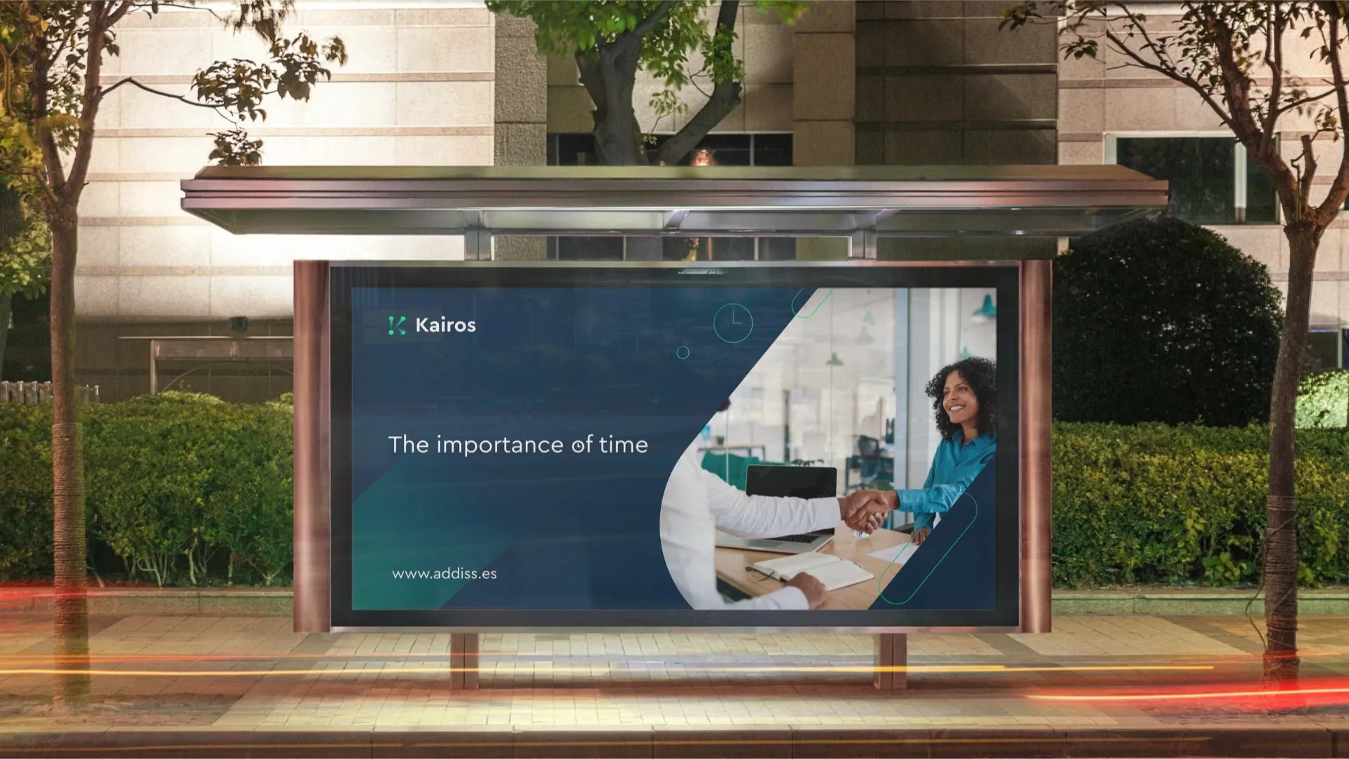  Gran panel publicitario en un paradero de autobús nocturno, mostrando un diseño de la marca 'Kairos' con la frase 'The importance of time' y una fotografía de dos profesionales dándose la mano en una oficina