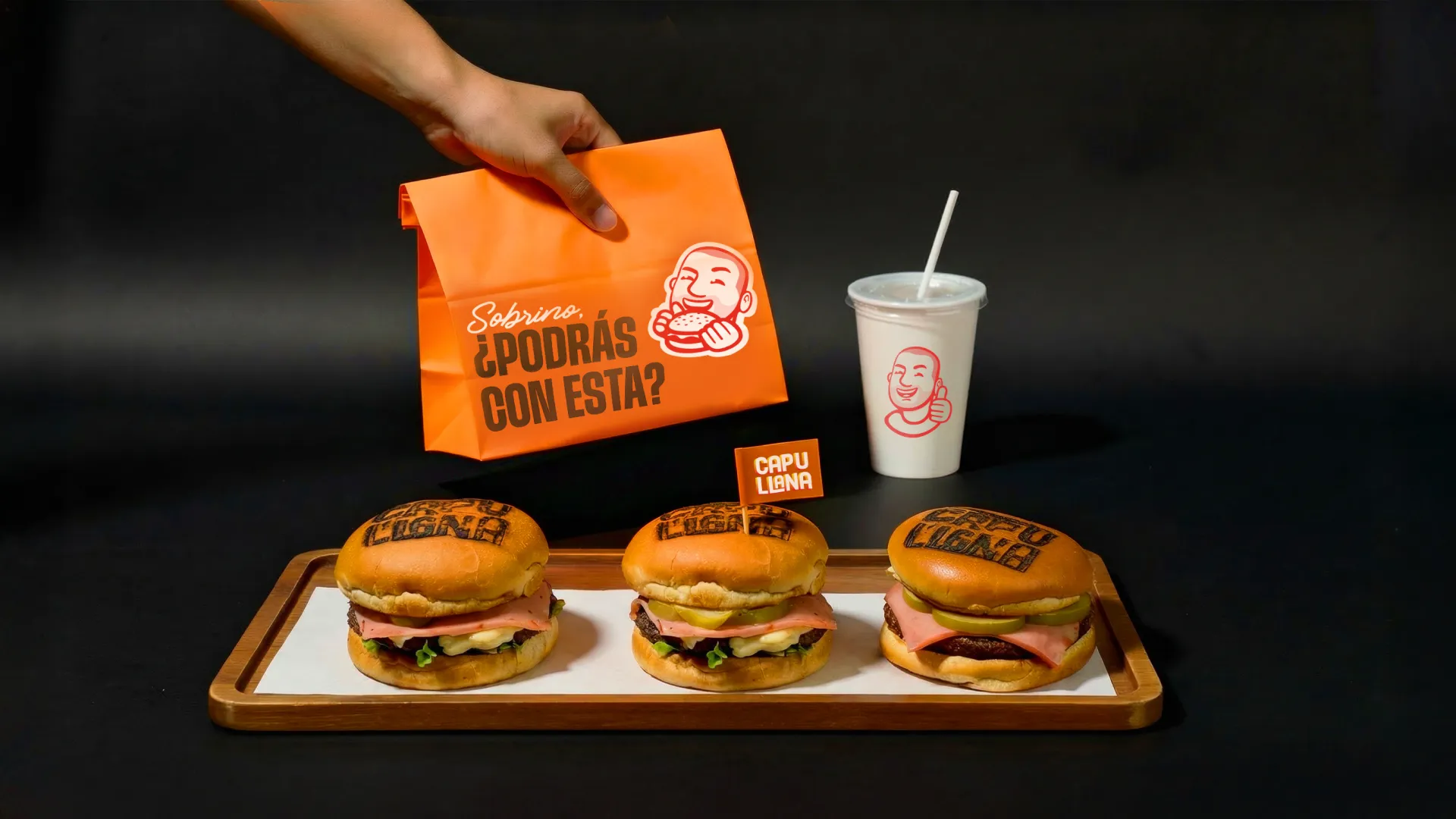 Mano sosteniendo una bolsa de papel naranja sobre una bandeja de madera con tres hamburguesas Capullana y un vaso blanco al lado