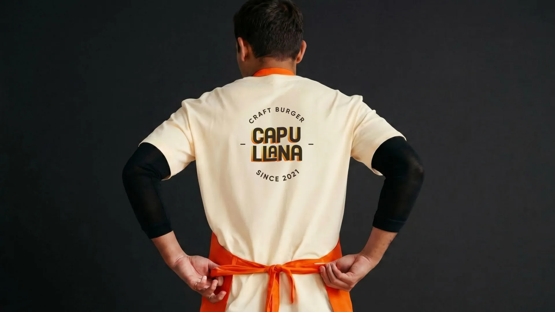 Persona de espaldas con camiseta clara de Capullana Craft Burger atándose un mandil naranja sobre fondo negro